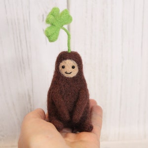 May include: A brown felt creature with a smiling face and a green four-leaf clover growing from its head.