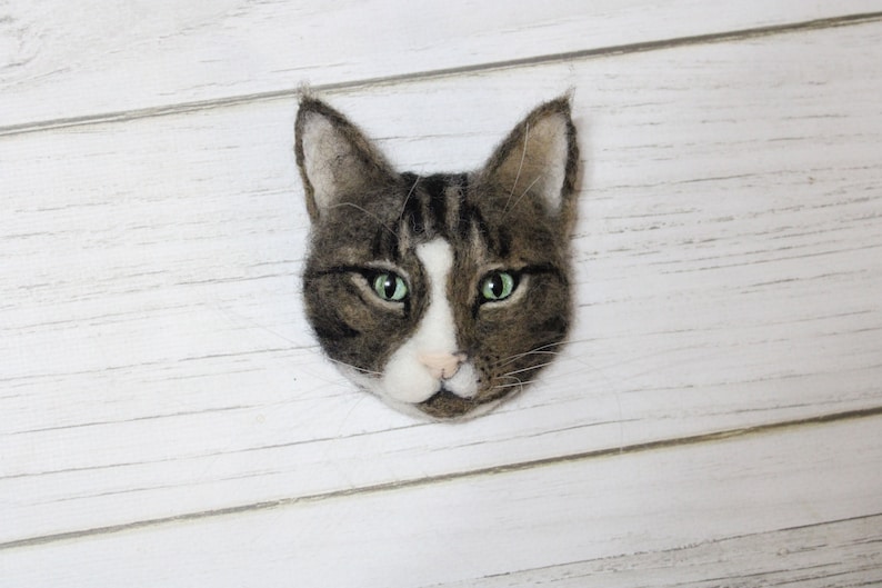 Puede incluir: Una cabeza de gato de fieltro con pelo marr&oacute;n y blanco y ojos verdes. El gato est&aacute; mirando directamente a la c&aacute;mara.