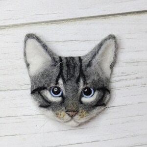Puede incluir: Una cabeza de gato de fieltro gris y blanco con ojos azules. El gato tiene rayas negras en la cara y una nariz rosa.