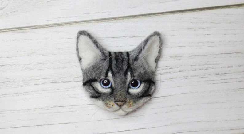 Puede incluir: Una cabeza de gato de fieltro gris y blanco con ojos azules. El gato tiene rayas negras en la cara y una nariz rosa.