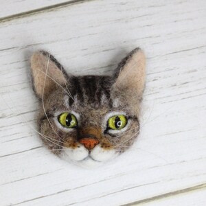 Puede incluir: Un broche de fieltro de un gato con un dise&ntilde;o realista. El gato tiene pelo marr&oacute;n y gris, ojos verdes y una nariz y barbilla blancas.