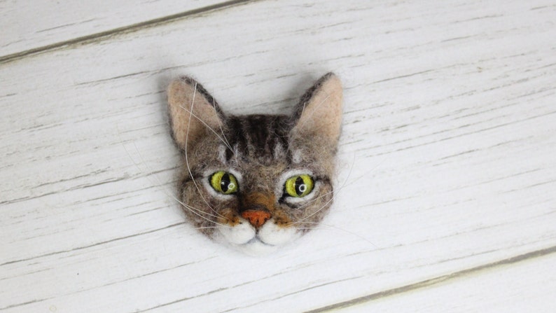 Puede incluir: Un broche de fieltro de un gato con un dise&ntilde;o realista. El gato tiene pelo marr&oacute;n y gris, ojos verdes y una nariz y barbilla blancas.