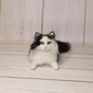 May include: A handmade, fluffy cat figurine with black and white fur. The cat has yellow eyes and is lying down. The figurine is made of a soft, textured material, and is set against a light wooden background.