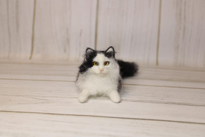 May include: A handmade, fluffy cat figurine with black and white fur. The cat has yellow eyes and is lying down. The figurine is made of a soft, textured material, and is set against a light wooden background.