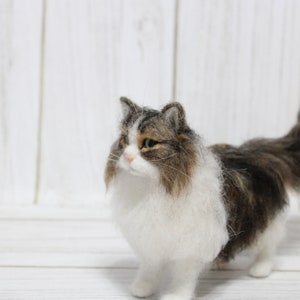 May include: A handmade, felted cat figurine with a fluffy, long-haired appearance. The cat has a white chest and belly, with brown and gray markings on its back and head. It stands on four legs, with a long, bushy tail. The cat's eyes are green.