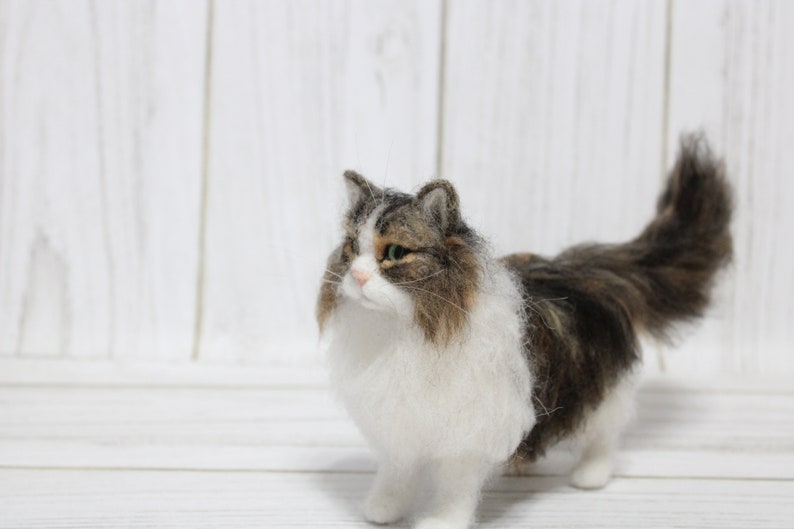 May include: A handmade, felted cat figurine with a fluffy, long-haired appearance. The cat has a white chest and belly, with brown and gray markings on its back and head. It stands on four legs, with a long, bushy tail. The cat's eyes are green.
