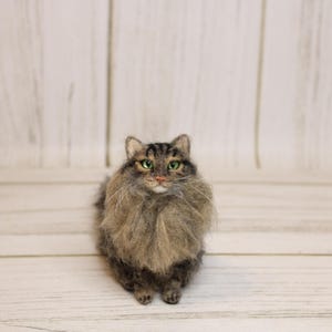 May include: A handcrafted, felted cat figurine with a fluffy, gray and brown coat. The cat has green eyes and a detailed face, standing on a light wood surface. The cat's fur is meticulously crafted, giving it a realistic appearance.
