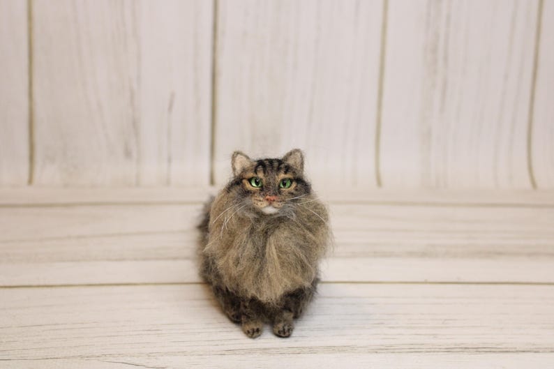 May include: A handcrafted, felted cat figurine with a fluffy, gray and brown coat. The cat has green eyes and a detailed face, standing on a light wood surface. The cat's fur is meticulously crafted, giving it a realistic appearance.