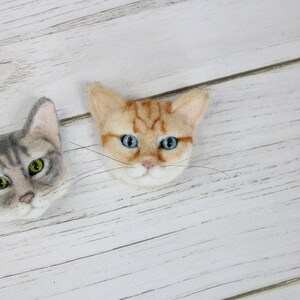 Puede incluir: Dos adornos de cabeza de gato de fieltro, uno gris y blanco con ojos verdes, el otro naranja y blanco con ojos azules.
