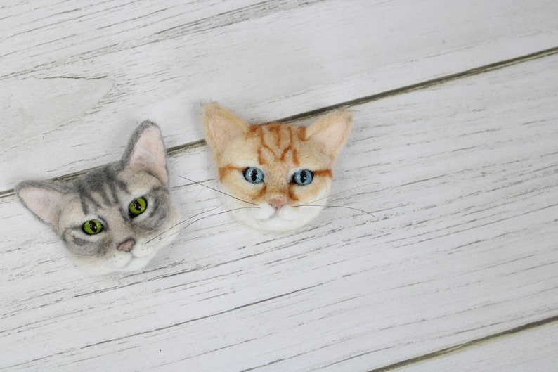 Puede incluir: Dos adornos de cabeza de gato de fieltro, uno gris y blanco con ojos verdes, el otro naranja y blanco con ojos azules.