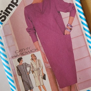 Può includere: Modello di cucito Simplicity 6105 con una donna in un abito viola. Il modello include illustrazioni di diversi stili di abiti e il testo "Cathy Hardwick". Il modello è circondato da un bordo a righe blu e bianche.