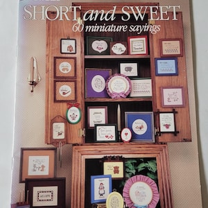 May include: A vintage cross-stitch pattern booklet titled "Short and Sweet" with 60 miniature sayings. The cover features a wooden cabinet with various cross-stitch designs in frames.