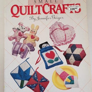 Puede incluir: Un libro titulado "Small Quiltcrafts" de Jennifer Geiger, que presenta una variedad de pequeños proyectos de acolchado, incluyendo una almohada en forma de corazón, un conejito, un cordero, una bolsa con forma de flor, un bloque de acolchado, un posavasos y un anillo para servilletas. La portada del libro es rosa y blanca con una etiqueta de precio de 14,95 €.