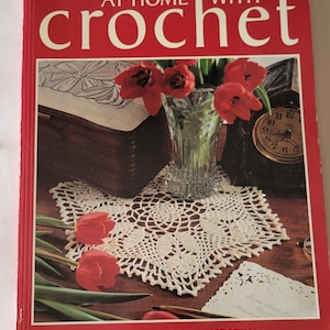Könnte beinhalten: Ein Buch mit dem Titel "At Home With Crochet" mit einem roten Einband und einer weißen gehäkelten Tischdecke auf der Vorderseite. Die Tischdecke ist von roten Tulpen und einer Uhr umgeben.