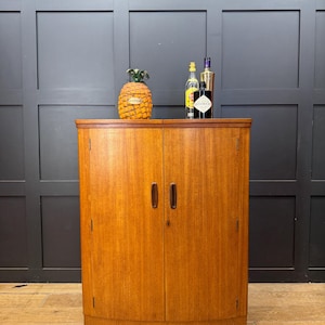 May include: A light brown wooden cabinet with two doors, featuring vertical wood grain, dark brown oval handles, and a curved front. On top sits a yellow pineapple-shaped container and three glass bottles of various sizes and labels.