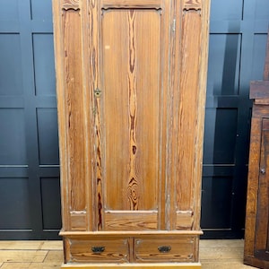 May include: A tall, light brown wooden wardrobe with two drawers at the bottom. The wardrobe has a single door and decorative vertical panels. The wood grain is visible throughout. The text "MAJOR P WILTSHIRE." is printed on the bottom.