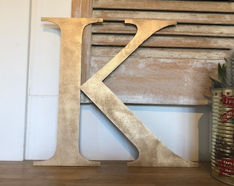 May include: A large, gold-colored wooden letter "K" stands on a wooden surface. The letter has a textured, metallic finish. The background includes a white wall, a wooden shutter, and a decorative tin.