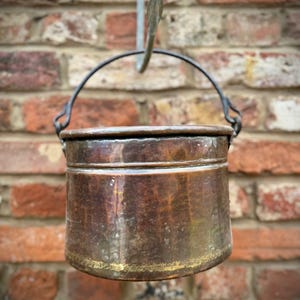May include: An aged copper pot with a black handle, suspended from a metal hook against a brick wall. The pot shows signs of wear, with a golden band at the base. It appears to be a small cooking pot, likely used in the past.