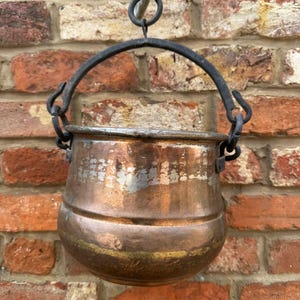 May include: An antique copper pot with a black iron handle and hanging ring. The pot has a rounded body and is suspended against a weathered brick wall. The copper has a warm, aged patina, showing signs of use and age. The pot is likely of a small size.