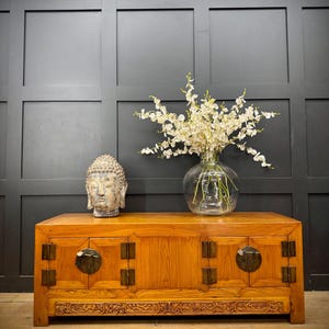 May include: A low rectangular wooden cabinet with a natural grain finish, featuring two sets of double doors with dark circular metal hardware and hinges. A stone Buddha head sculpture and a clear glass vase with white flowers sit on top.