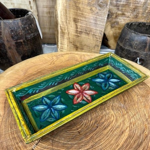 May include: A rectangular wooden tray with a green background and a yellow border. The tray is decorated with three painted flowers in shades of blue, red, and pink.