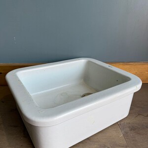 May include: A white rectangular porcelain sink with a rounded edge. The sink features a drain hole and is set against a blue-grey wall and a wooden floor. This classic sink design is suitable for a bathroom or utility room.