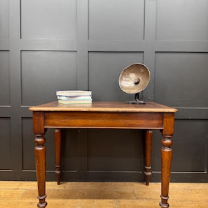 May include: A rectangular wooden table with four turned legs and a smooth top surface. A stack of three books with white and blue covers sits on the left side, and a metallic, bowl-shaped desk lamp with a black base sits on the right side.
