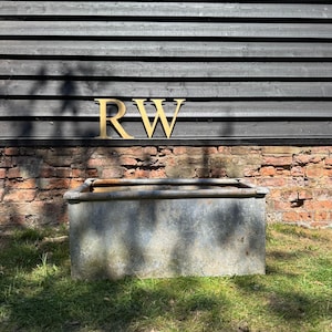 May include: A galvanised metal trough on grass, set against a brick wall and a dark wooden backdrop. Gold-coloured letters "RW" are displayed against the dark wood.
