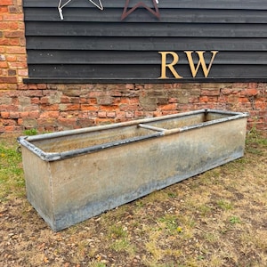 May include: A long, rectangular galvanised metal trough with a weathered, silver-grey finish. The container features a reinforced rim and a central metal support bar spanning the width of the basin, showing signs of oxidation and outdoor use.