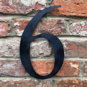 May include: Black metal number six, attached to a brick wall. The number is large and has a slightly curved design.