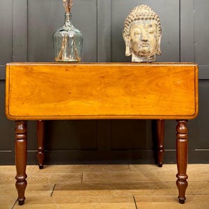 May include: A wooden drop leaf table with a light brown finish and turned legs. The table is closed and has a smooth surface.