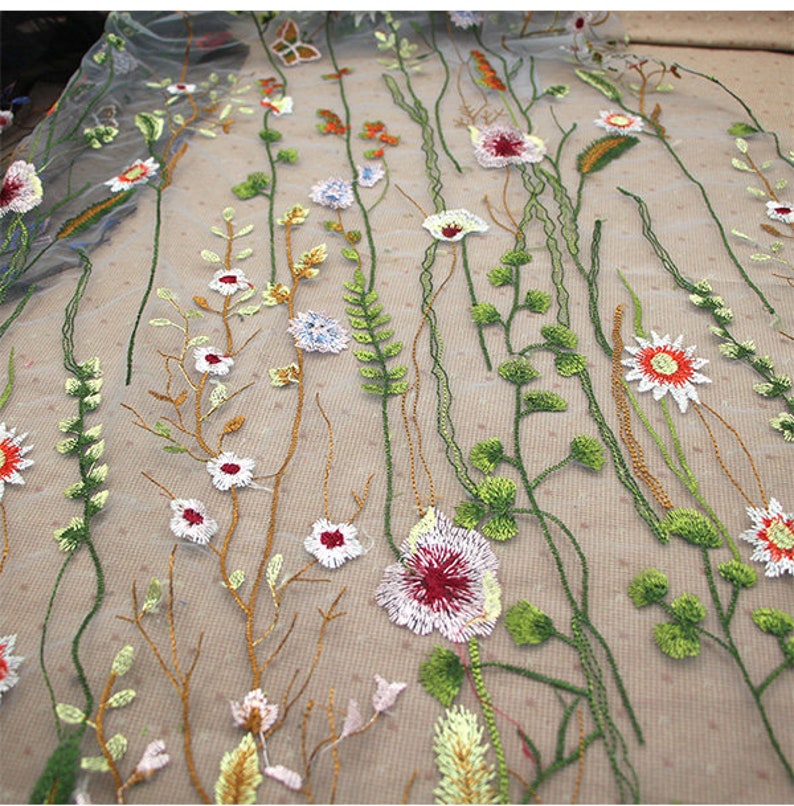 K&ouml;nnte beinhalten: Nahaufnahme eines bestickten floralen Stoffes. Der Stoff weist ein zartes Muster aus verschiedenen Blumen und Bl&auml;ttern in Wei&szlig;, Rot, Orange und Gr&uuml;n mit braunen Stielen und Bl&auml;ttern auf. Der Hintergrund ist hellbeige.