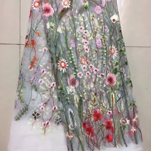 K&ouml;nnte beinhalten: Transparentes besticktes Gewebe mit floralem Muster. Die Stickerei zeigt rosa, rote und wei&szlig;e Blumen mit gr&uuml;nen Stielen und Bl&auml;ttern. Der Stoff hat unten einen wei&szlig;en Rand.