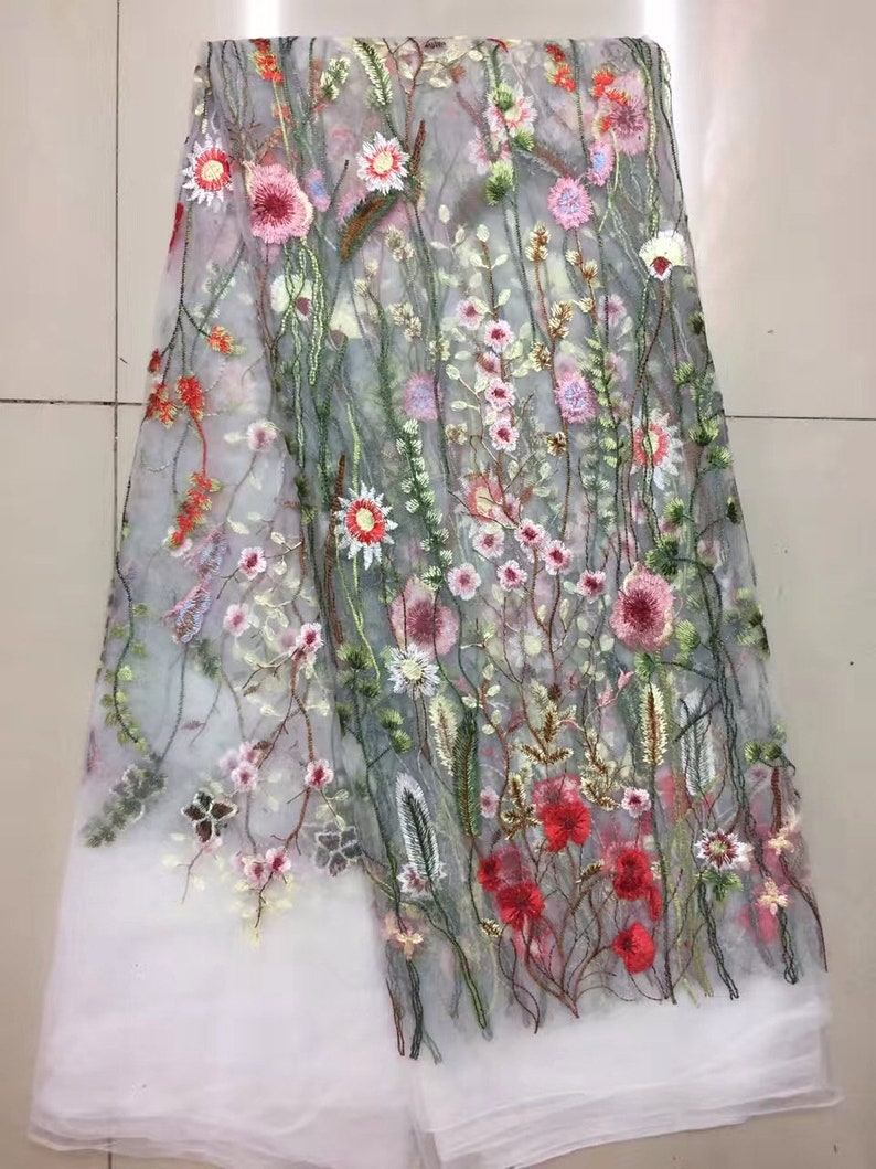 K&ouml;nnte beinhalten: Transparentes besticktes Gewebe mit floralem Muster. Die Stickerei zeigt rosa, rote und wei&szlig;e Blumen mit gr&uuml;nen Stielen und Bl&auml;ttern. Der Stoff hat unten einen wei&szlig;en Rand.