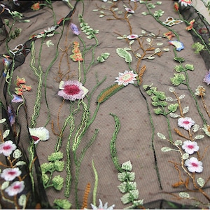 K&ouml;nnte beinhalten: Nahaufnahme eines schwarzen, transparenten Stoffes mit gesticktem Blumendesign. Die Stickerei zeigt eine Vielzahl farbenfroher Blumen, darunter rosa, wei&szlig; und lila, mit gr&uuml;nen Stielen und Bl&auml;ttern. Der Stoff hat ein gepunktetes Muster.
