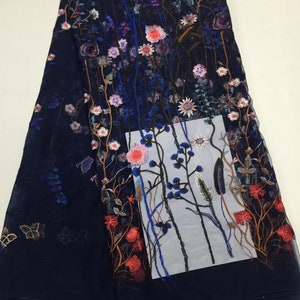 K&ouml;nnte beinhalten: Marineblau bestickter Stoff mit floralem Design. Der Stoff weist ein detailliertes Muster aus bunten Blumen auf, darunter rosa, rote und wei&szlig;e Bl&uuml;ten, mit gr&uuml;nen Bl&auml;ttern und Stielen. Auch Schmetterlinge sind auf dem Stoff gestickt.