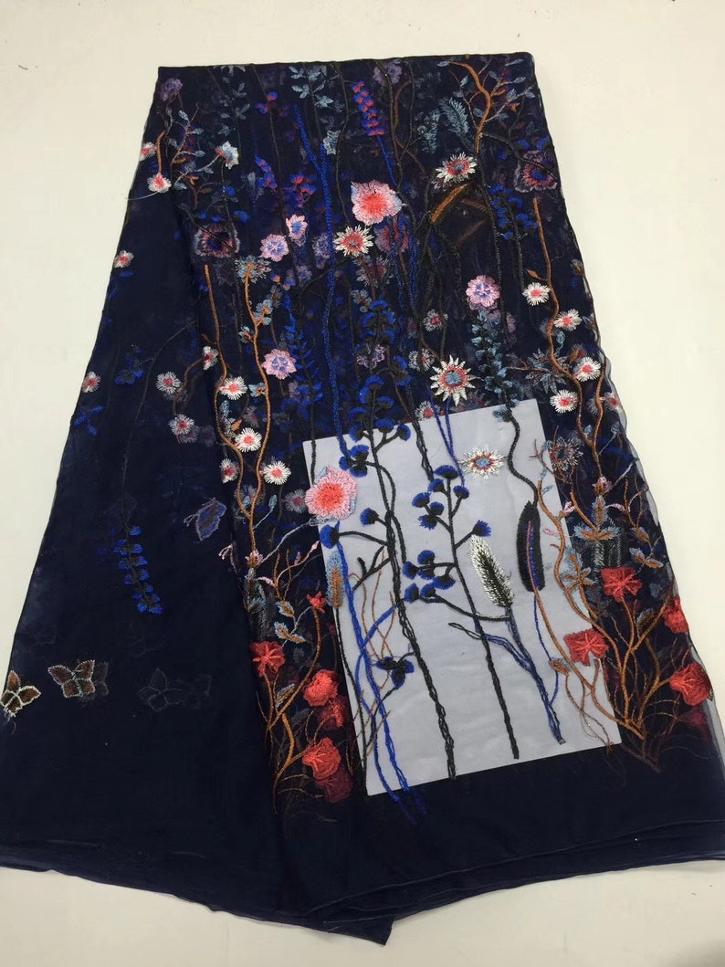 K&ouml;nnte beinhalten: Marineblau bestickter Stoff mit floralem Design. Der Stoff weist ein detailliertes Muster aus bunten Blumen auf, darunter rosa, rote und wei&szlig;e Bl&uuml;ten, mit gr&uuml;nen Bl&auml;ttern und Stielen. Auch Schmetterlinge sind auf dem Stoff gestickt.