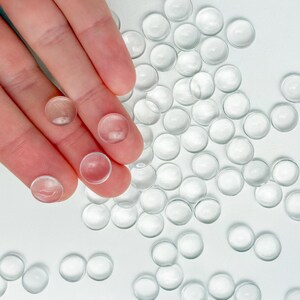 May include: Clear, round glass gems scattered on a white surface. Several gems are held by a hand. The gems are transparent and have a slightly domed shape. These decorative glass gems are suitable for crafts and home decor.