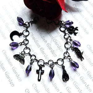 May include: A black charm bracelet with various charms, including a crescent moon, cross, and hand. The bracelet features purple teardrop-shaped beads and is set against a background with the text "Obsidian Garden".