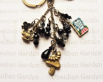 May include: A gold-tone keychain with a charm featuring a book with the text "Through the Looking Glass". The keychain also has black beads, a teapot charm, a mushroom charm, and a spade charm.