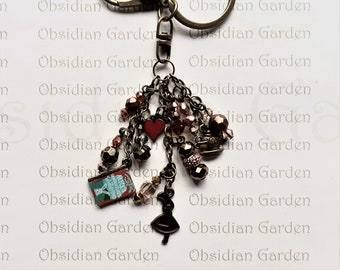 May include: A bronze key chain with a charm featuring a book, a heart, a teacup, and a small figure of Alice in Wonderland. The charm is adorned with beads and chains.