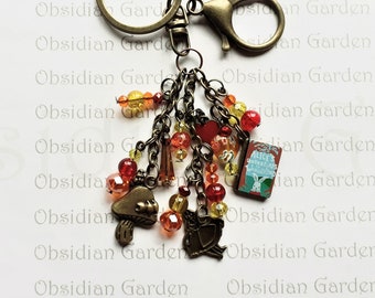 May include: A bronze key chain with a charm featuring a miniature book with the title "Alice's Adventures in Wonderland". The charm is attached to a chain with multiple charms, including a mushroom, a bird, and beads in shades of red, orange, and yellow.