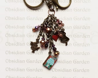 May include: A bronze key chain with a charm featuring a book with the text "Alice's Adventures in Wonderland". The charm is attached to a chain with several charms, including a red heart, a bell, and a bird. The key chain is a great gift for fans of Alice in Wonderland.