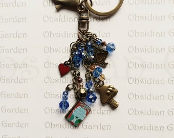 May include: A bronze key chain with a variety of charms, including a red heart, a blue crystal, a mushroom, a book with the title "Alice's Adventures in Wonderland", and a small golden bell. The key chain is attached to a split ring.