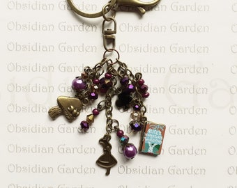 May include: A bronze-colored keychain with a charm featuring a purple and brown beaded design, a mushroom charm, a miniature Alice in Wonderland book charm, and a miniature Alice charm. The keychain has a split ring and a lobster clasp.