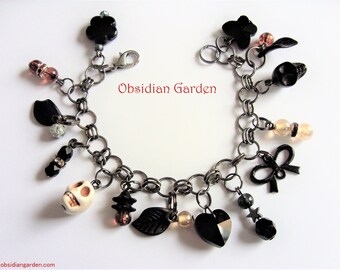 May include: A black chain bracelet with multiple charms, including a skull, a heart, a bow, leaves, and beads. The bracelet is adorned with black, white, and silver accents. The text "Obsidian Garden" is visible in the image.