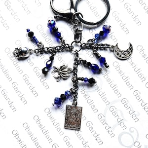 May include: A silver-coloured keyring featuring a selection of charms, such as a skull, spider, crescent moon, and a rectangular pendant. The charms are linked by dark chains and embellished with blue and black beads. The keyring has a lobster clasp and a split ring.