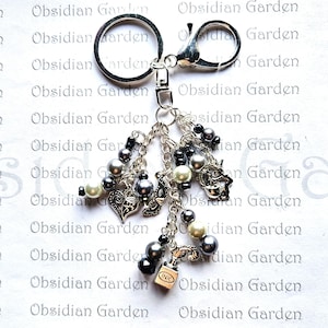 May include: A silver charm keychain with a variety of charms, including a heart, a bird, a bottle of ink, and beads. The charms are attached to a silver chain and a silver key ring.