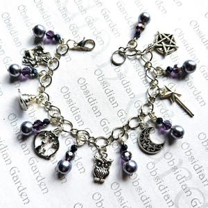May include: A silver charm bracelet featuring various charms, including a pentagram, moon, owl, and wand. The bracelet also has gray pearl beads and purple crystal accents. The text "Obsidian Garden" is printed on the background.