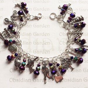 Op de afbeelding: Zilveren bedelarmband met diverse bedels, waaronder een medisch kruis, een granaat, een bijl en een boom. De armband heeft ook paarse en iriserende kralen. De tekst "Obsidian Garden" is zichtbaar op de achtergrond.
